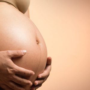 Barriga de mujer embarazada con manos sosteniéndola, fondo de color neutro.