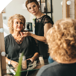 Mujer con cabello rizado recibe maquillaje en un salón, reflejada en un espejo.