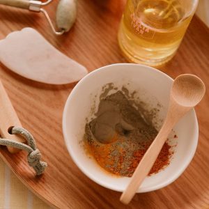 Vaso con mezcla de polvo y especias, rodillo facial y aceite en una bandeja de madera.